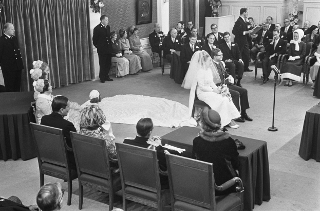 Schwarzes und weißes Foto einer Hochzeitszeremonie mit Menschen, die sitzen und stehen, einem Mann mit Mikrofon, einer Frau mit einem Strauß Blumen, einem Podium, Blumengestecken, einem Vorhang, einem gerahmten Foto an der Wand und einer Tür.