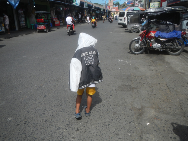 Ein junger Junge geht eine Straße entlang, die von Motorrädern und anderen Fahrzeugen gesäumt ist, mit Gebäuden, Namensschildern, Bäumen, Pfählen und einem klaren blauen Himmel im Hintergrund.