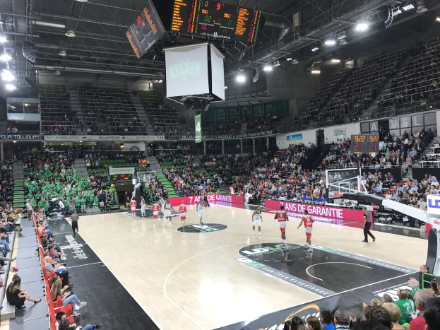 Basketballspiel in einer großen Arena mit Publikum, Anzeigetafeln mit "Asvel vs Galatasaray" und Überdachung mit Bildschirm.