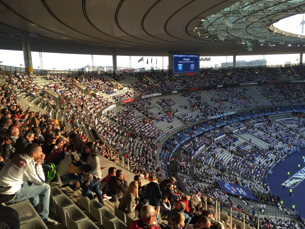 Große Menschenmenge im Allianz Arena Stadion bei einem Fußballspiel sitzend, mit einer Bühne rechts, Fahnen, Stangen, einem Bildschirm und einem sichtbaren Himmel im Hintergrund.