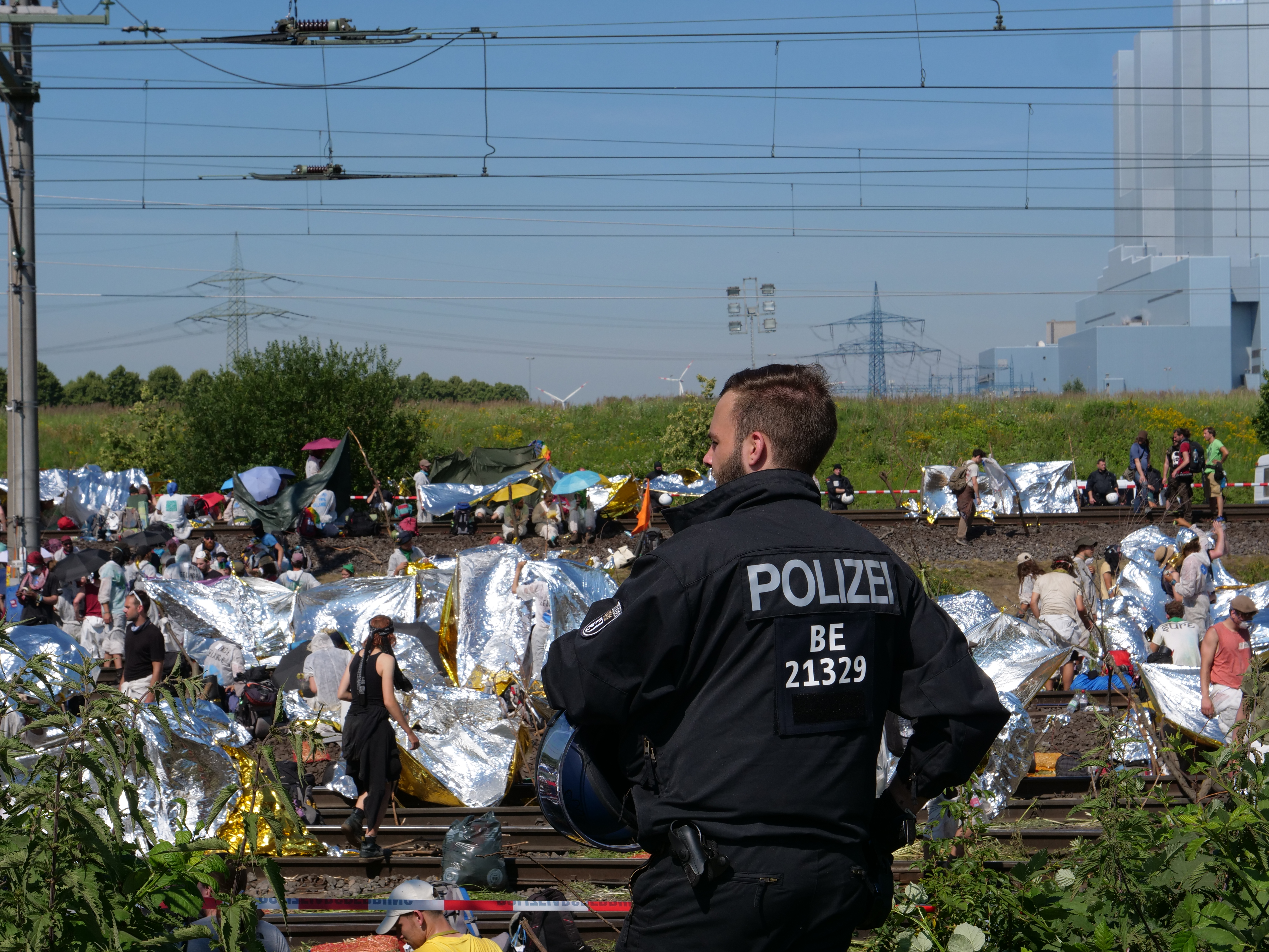 Ein Polizist in schwarzer Jacke steht vor einer Menschenmenge auf den Schienen, mit Bäumen, einem blauen und weißen Himmel und Strommasten im Hintergrund.