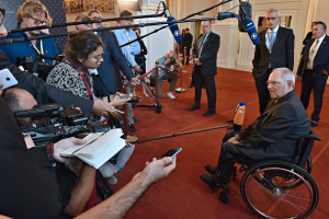 Ein Mann im Rollstuhl umgeben von Reportern mit Kameras, Handys und Mikrofonen mit Ständern, einem Banner, Scheinwerfern und einer Wand im Hintergrund.