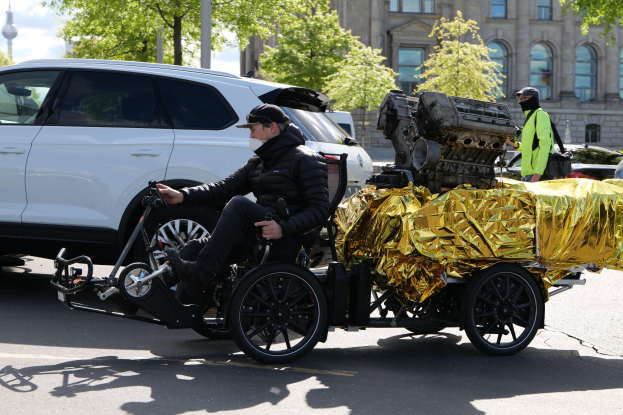 Ein Mann im Rollstuhl mit einem großen Motor an dessen Rückseite, umgeben von Fahrzeugen auf einer Straße mit Bäumen, Gebäuden und einem klaren blauen Himmel im Hintergrund.