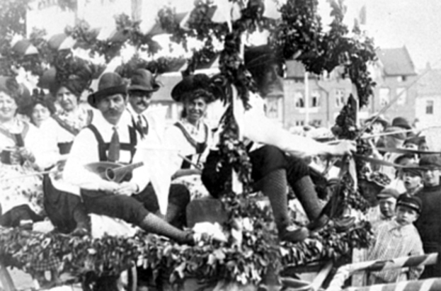 Schwarzes und weißes Bild eines Paradewagens mit Menschen in traditioneller bayrischer Tracht, einige spielen Musikinstrumente, mit Blattdekoration vor einem Hintergrund aus Gebäuden und einem klaren Himmel.