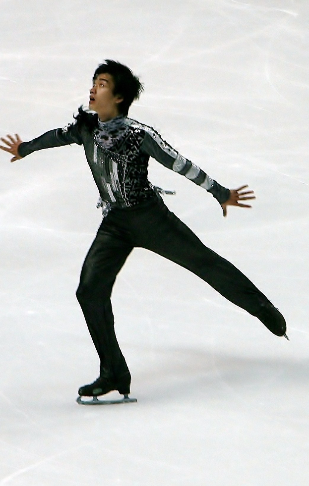 Ein männlicher Eiskunstläufer in einem schwarzen Kleid führt eine Routine auf einem Eisstadion durch und gleitet dabei elegant mit synchronisierten Arm- und Beinbewegungen.