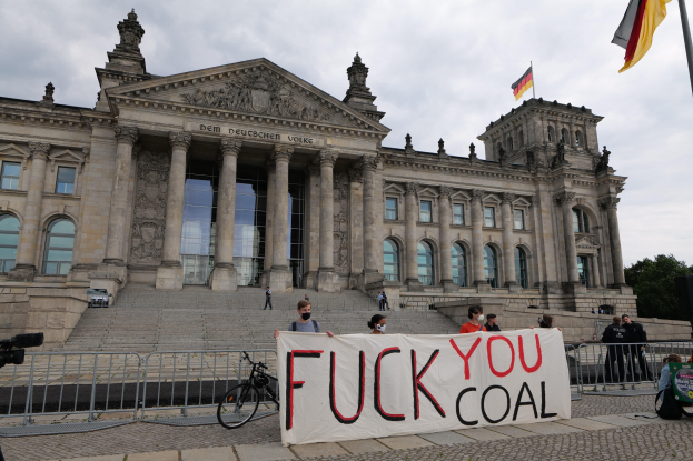 Protestierende mit einem "Fuck You Coal"-Schild vor dem Reichstaggebäude in Berlin, Deutschland, mit seinen architektonischen Details, einem Fahrrad, Bäumen, einer Flagge und einem bewölkten Himmel.