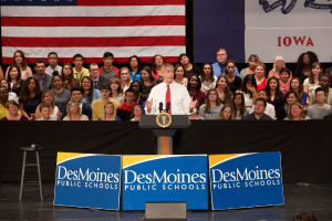 Ein Mann in einem weißen Hemd und roter Krawatte steht an einem Rednerpult mit drei Texttafeln, einem Hocker mit Mikrofon und einer sitzenden Menge im Hintergrund, mit einer Flagge und zusätzlichem Text.