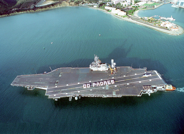 Das Flugzeugträger USS George H W Bush CVN-68 ist zu sehen, wie es im Pazifischen Ozean segelt, mit mehreren Schiffen und Booten auf seinem Deck und Gebäuden, Bäumen und Bergen im Hintergrund.