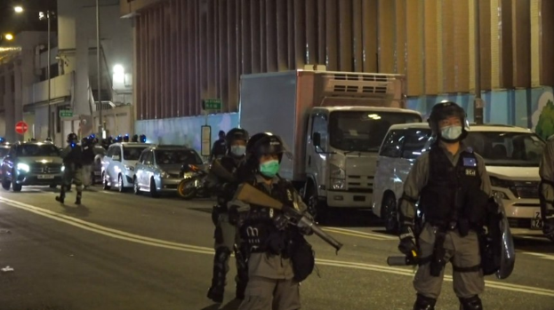 Eine Gruppe von Polizisten in Helmen, Handschuhen und Gesichtsmasken steht an einer Straße im Dunkeln, einige halten Waffen, mit Fahrzeugen, Laternen, Schildern und Gebäuden im Hintergrund.