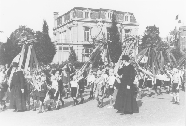 Schwarzes und weißes Bild einer Gruppe von Menschen, die eine Straße entlanggehen und Fahnen schwenken, mit Hüten, Maibaum, Bäumen, Gebäuden und Himmel.