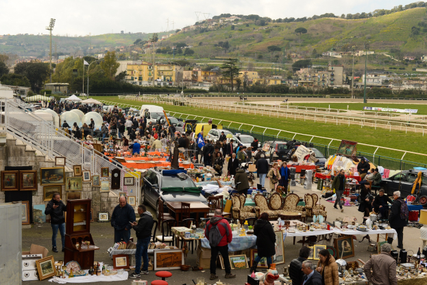 Große Gruppe von Menschen auf einem Flohmarkt mit Tischen, die Gegenstände wie Fotorahmen und Stühle anbieten, umgeben von geparkten Fahrzeugen, Geländern, Treppen, Bäumen, Gebäuden, Laternenpfählen, Hügeln und einem bewölkten Himmel.