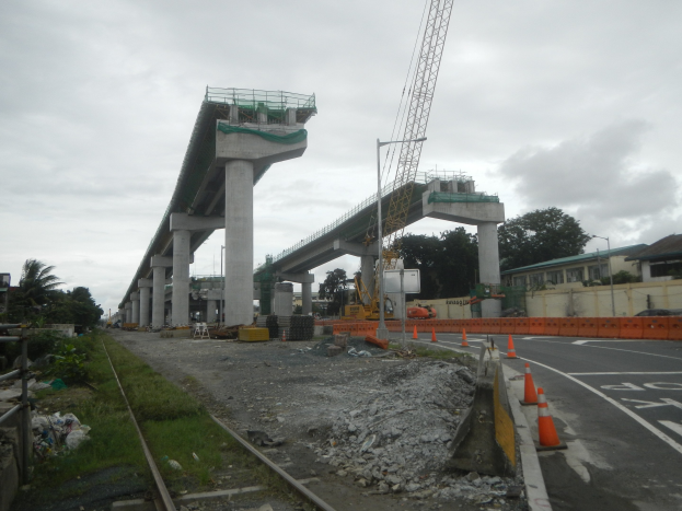 Eine Baustelle mit einer Brücke im Hintergrund, eine Straße mit Verkehrskegeln auf der rechten Seite, Steine und Gras am Boden, eine Bahnschiene auf der linken Seite, Bäume und Gebäude auf beiden Seiten der Straße und ein bewölkter Himmel.