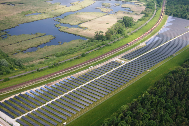 Luftaufnahme einer Solaränlage mit Paneelen, umgeben von Bäumen, Gras und Wasser, mit einem Zug auf einer näheren Eisenbahnstrecke.