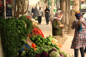 Ein belebter Gemüsemarkt mit verschiedenen Produkten wie Paprika, Tomaten und Kartoffeln auf der linken Seite, Menschen, die auf der rechten Seite stehen und gehen, und Bäume und Gebäude im Hintergrund.