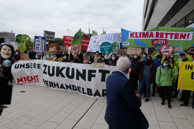 Eine Gruppe von Menschen, die gegen den Klimawandel protestieren und Schilder und Plakate in der Hand halten, vor einem Gebäude stehend, mit einer Person, die eine Kamera auf der linken Seite hält, und Bäumen, Pfählen und einem klaren blauen Himmel im Hintergrund.