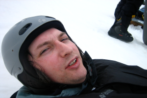 Ein Mann in einem Pullover und Helm steht im Schnee, mit den Beinen und Skiern einer anderen Person im oberen rechten Eck.