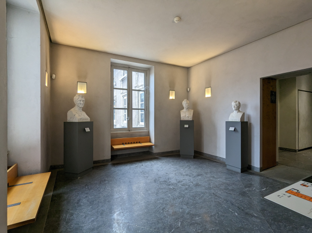 Ein Raum mit Skulpturen auf Sockeln, einer hölzernen Bank, einem Fenster, an der Wand montierten Lampen, einer Tür und einer Decke, wahrscheinlich im Museum of Fine Arts in Paris.