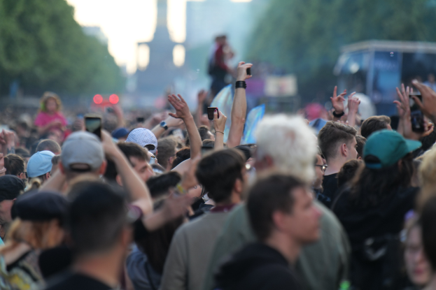 Menschenmenge auf einem Musikfestival mit erhobenen Händen, einige tragen Mützen und halten Telefone, vor einem unscharfen Hintergrund aus Bäumen, Gebäuden, Lichtern und einem Fahrzeug.
