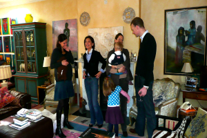 Eine Gruppe von Menschen steht in einem Raum mit Büchern, Stühlen, einem Telefon, Lampen, Schränken und Gemälden an der Wand.