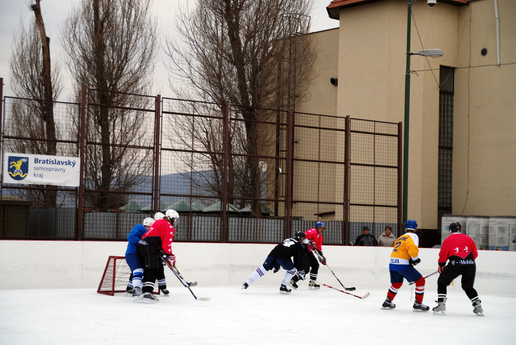 Menschen beim Eishockey auf einem Eisplatz mit Gebäuden, Bäumen, einer Straßenlaterne, einem Namensschild und Zäunen im Hintergrund unter dem Himmel.