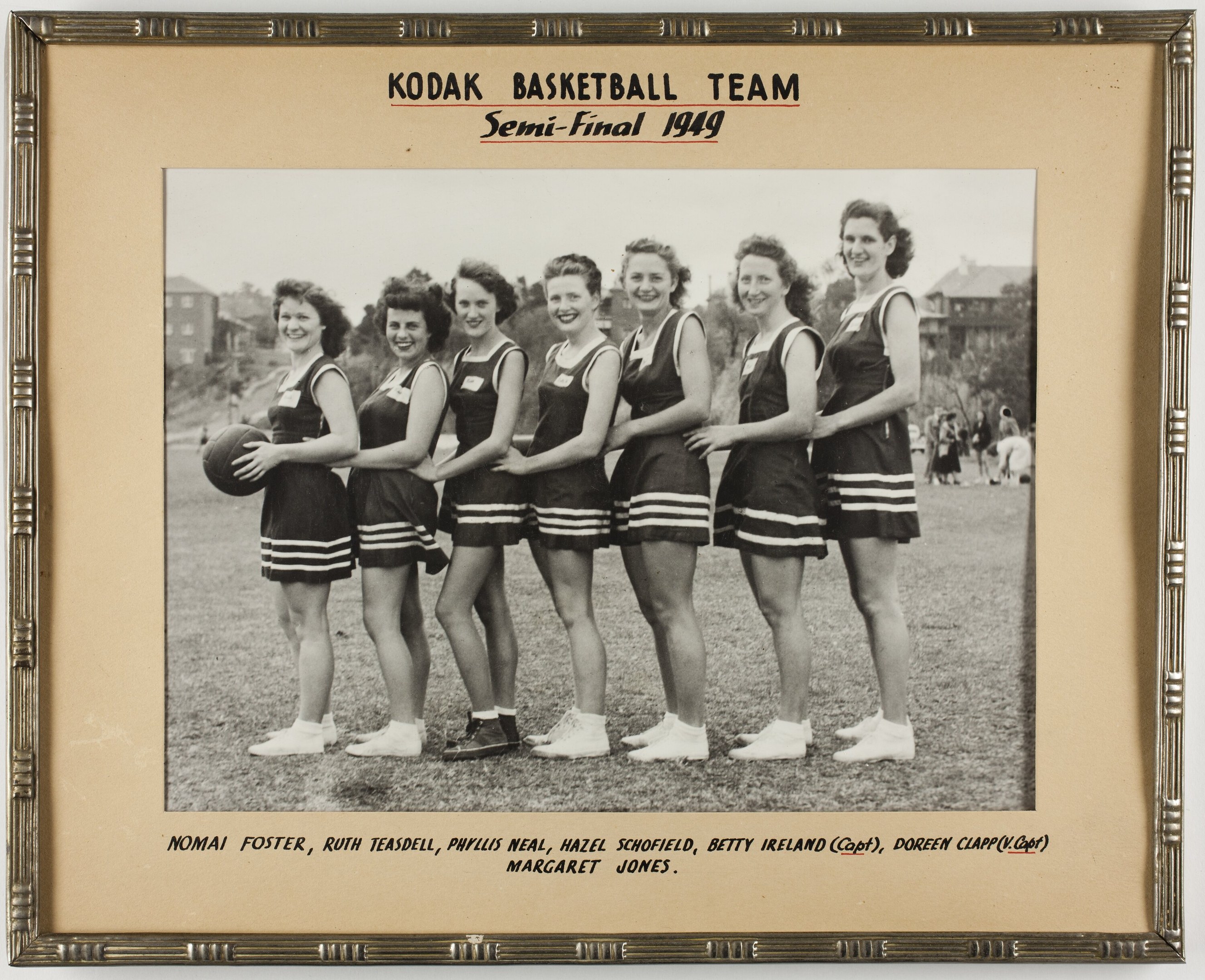 Schwarzes und weißes Foto einer Gruppe von Frauen der Kodak Basketball Mannschaft, die lächeln und einen Basketball halten, mit Zuschauern, Bäumen, Gebäuden und Himmel im Hintergrund.