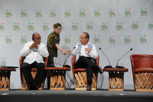 Zwei Männer sitzen auf Stuhl auf der Bühne, einer bekommt ein Papier von einer Frau an einem Schreibtisch mit zwei Mikrofonen, während ein anderer Person mit einem Headset das Papier ansieht; ein Tisch mit einem Glas ist zwischen den Männern, und eine Tafel mit Bildern ist im Hintergrund.