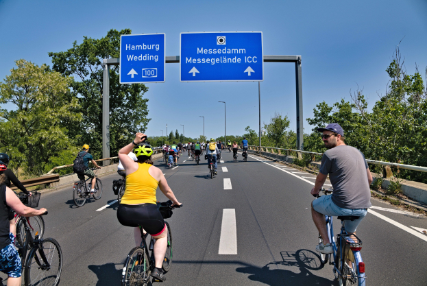 Eine Gruppe von Radfahrern in Helmen fährt eine Straße mit einer Begrenzung auf einer Seite und Bäumen auf der anderen entlang, unter einem klaren blauen Himmel mit Laternen im Hintergrund; ein Schild oben zeigt eine Radtour in Hamburg an.