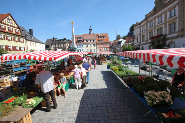 Ein belebter Markt im alten Stadtzentrum von Heidelberg mit Menschen, die spazieren gehen, auf Bänken sitzen und in der Nähe von Gemüsetischen unter Zelten stehen, vor dem Hintergrund von Gebäuden, Bäumen und einem klaren blauen Himmel.