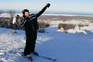 Eine Person auf einem Snowboard in der Mitte des Bildes, die auf Schnee mit Bäumen, Menschen, einem Pfahl, Wasser und Himmel im Hintergrund Ski fährt.