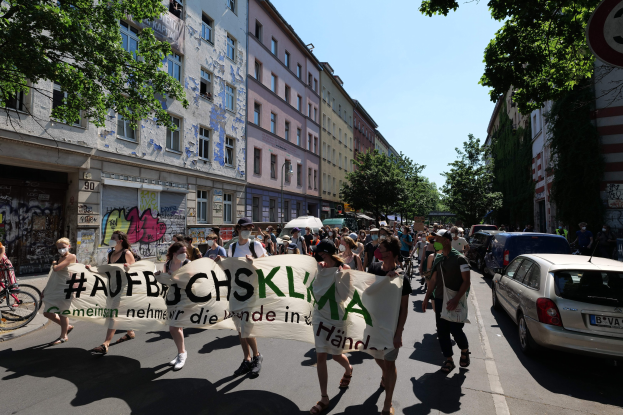 Eine Gruppe von Menschen, die eine Straße entlanggehen und ein Transparent halten, mit parkenden Autos auf der rechten Seite, einem Fahrradfahrer auf der linken Seite, Gebäuden und Bäumen im Hintergrund und einer Protestandrohung in Berlin.