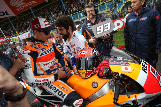Eine Gruppe von Menschen um ein Motorrad auf einer Rennstrecke versammelt, mit einer Person in einem weißen, orangen und roten Kleid und Helm in der Mitte, einer anderen Person mit einem Schirm links und Zuschauern im Hintergrund.