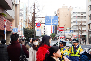 Eine volle Szene mit Menschen in verschiedenen Kleidern und Mützen, einer Handtasche, Haarsträhnen, einer Platte, einem Pfahl, Bäumen, Gebäuden mit Fenstern, einer Straße mit einer weißen Linie und einem weißen Himmel.