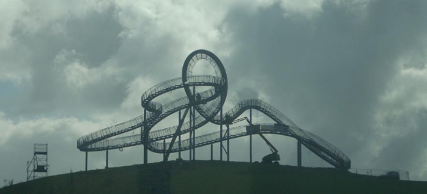 Eine Achterbahn auf dem Höhepunkt eines Hügels unter einem bewölkten Himmel, mit umgebenen Stützpfählen und Strukturen.