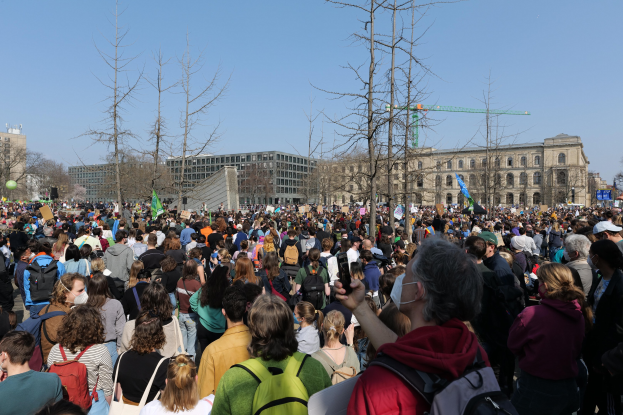 Große Menschenmenge mit Schildern und Taschen vor einem Gebäude mit Bäumen und einem klaren Himmel, was auf eine Klimaprotest hingedeutet.