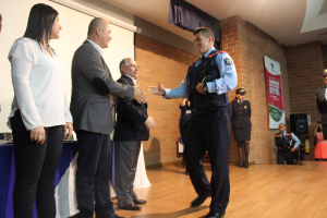 Eine Gruppe von Menschen auf einer B√ľhne, einer in Uniform mit einer M√ľtze, eine Frau in einem weißen Hemd, mit einem Banner, Lautsprecher, Vorh√§ngen, Tisch und Bildschirm im Hintergrund.