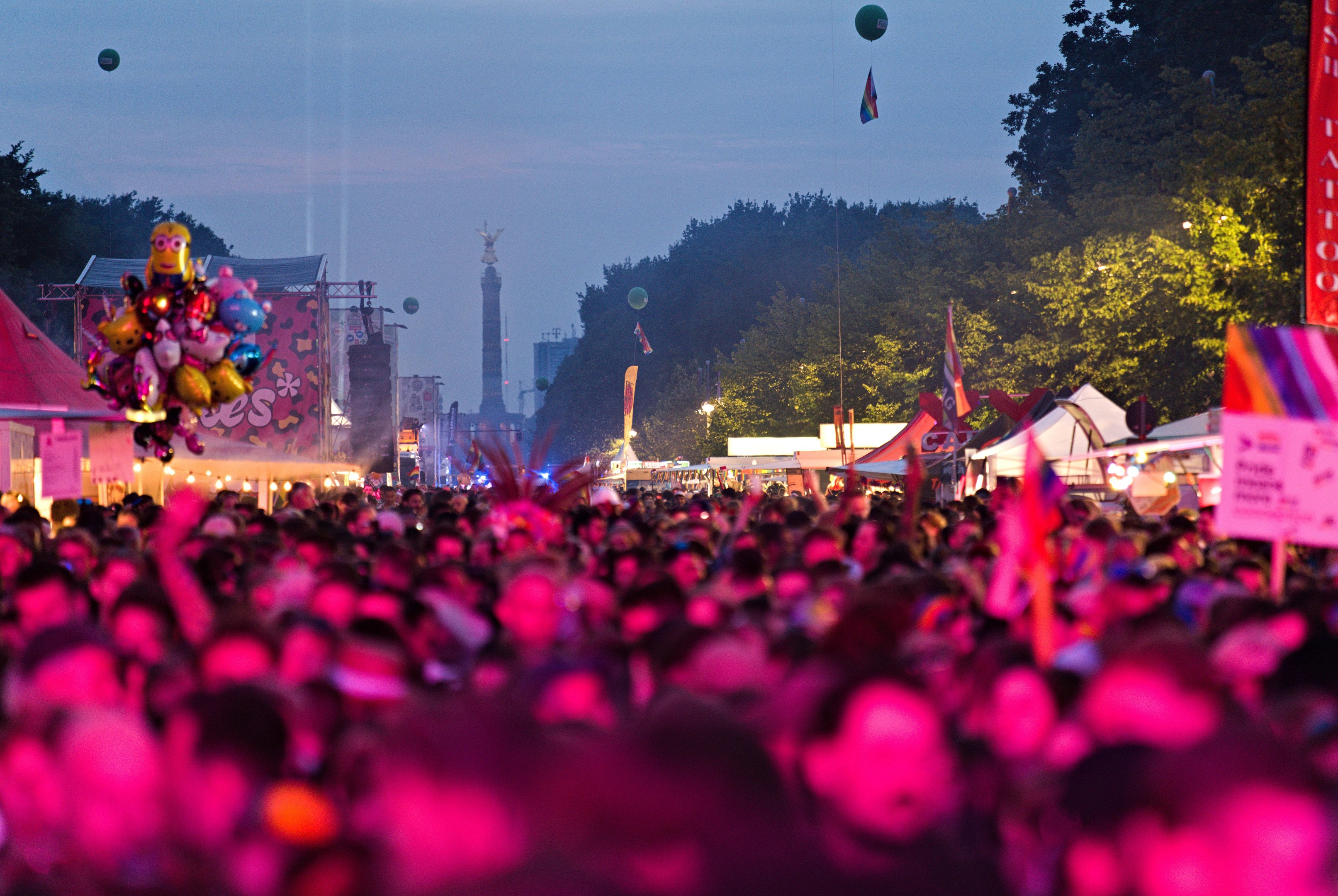 Eine große Menge auf einem nächtlichen Musikfestival mit bunten Lichtern, Ballons, Zelten, Bannern, Fahnen, Bäumen, einem Turm und einem sternenklaren Himmel im Hintergrund.