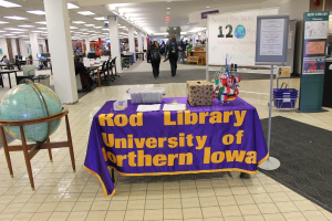 Messestand mit einem Tisch, auf dem Flaggen verschiedener Länder, eine Box, Papiere und eine Fahne zu sehen sind, sowie eine Weltkugel links, eine Anschlagtafel und Menschen rechts und zusätzliche Tische und Stühle im Hintergrund.