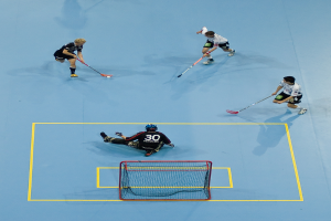 Drei Personen in Sportkleidung spielen Hockey, wobei eine Person rechts mit einem Hockeystock einen Ball schlägt und eine Person hinter einem Netz auf dem Boden sitzt.