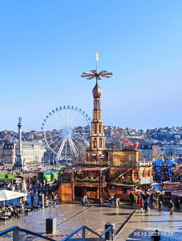 Ein belebter Stadtplatz mit einem zentralen Riesenrad, umgeben von Gebäuden, Bäumen und Menschen unter einem klaren blauen Himmel, mit Zelten und Geländern sichtbar.