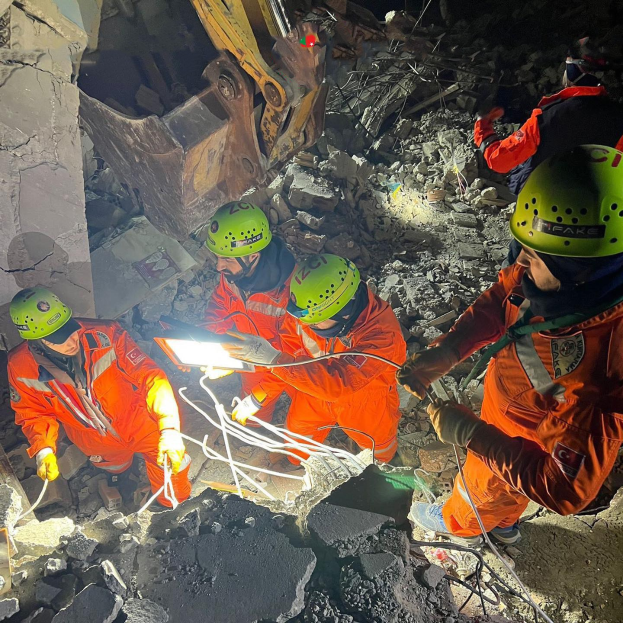 Eine Gruppe von Menschen in orangen Uniformen und grünen Helmen bei der Arbeit an Schutt mit einem Kran im Hintergrund während eines Einsatzes nach dem Einstürzen eines Gebäudes.