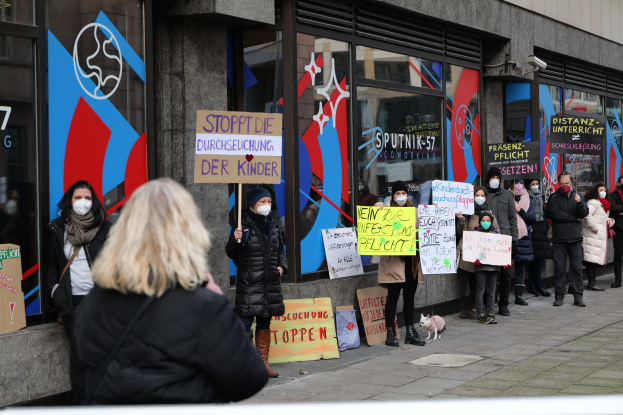 Eine Gruppe von Menschen mit Masken und Schildern protestiert vor einem Gebäude mit Glaswänden und -türen, mit zusätzlichem Protestmaterial und einem Hund.