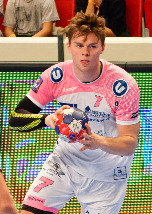 Ein Mann in einem rosa Hemd hält einen Fußball, während eine andere Person links zu sehen ist, mit einigen sitzenden Personen im Hintergrund.