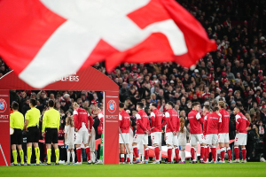 Eine Gruppe von Menschen auf einem Fußballfeld mit einer roten und weißen Flagge im Vordergrund, einem Bogen mit Text im Hintergrund und einer großen Menschenmenge im Stadion.