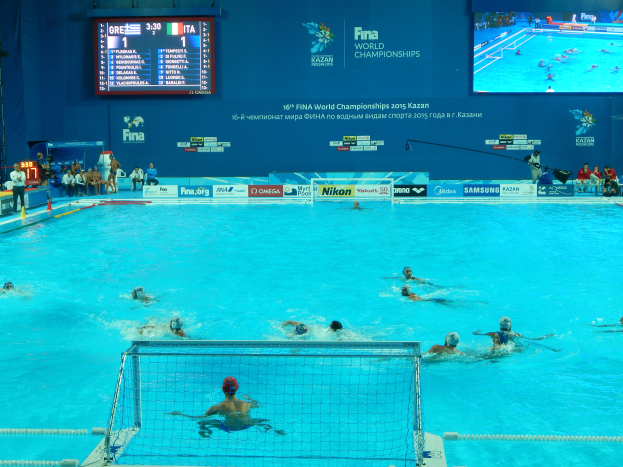Eine Gruppe von Menschen spielt Wasserball in einem großen Pool mit einem Netz in der Mitte, Zuschauer auf der linken Seite und ein Bildschirm mit Text und Logos im Hintergrund bei den 2016 FINA-Weltmeisterschaften in Kazan.