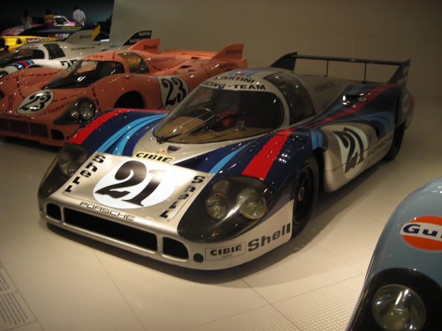 Ein Porsche 917K Rennwagen von Le Mans in einem Museum neben anderen Sportwagen ausgestellt, mit Menschen, die zuschauen, und einem Textschild auf der linken Seite.