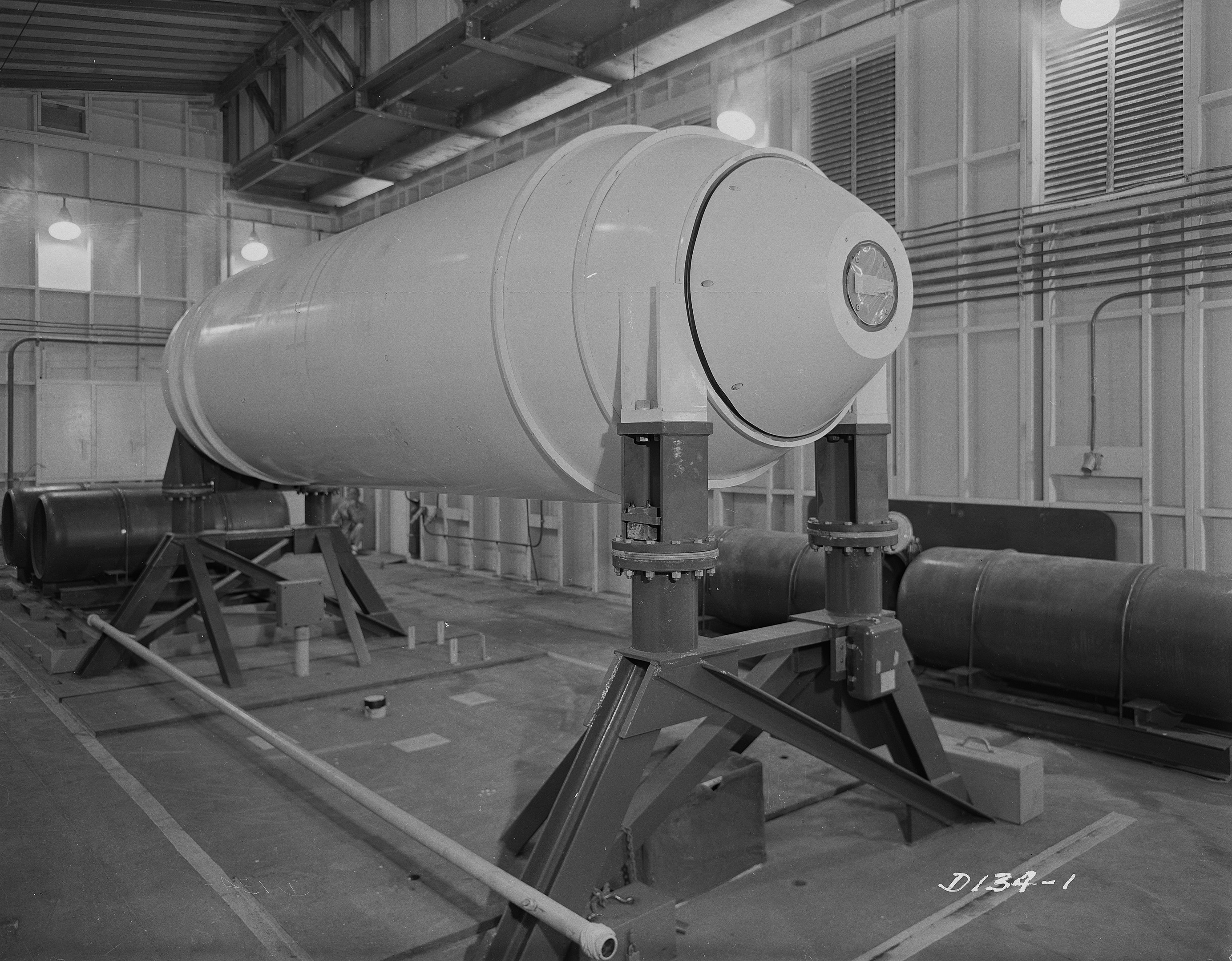 Großer zylindrischer Wasserstoffbomben auf einem Ständer in einer industriellen Fabrikumgebung mit Rohren, Lampen und einer Wand im Hintergrund.