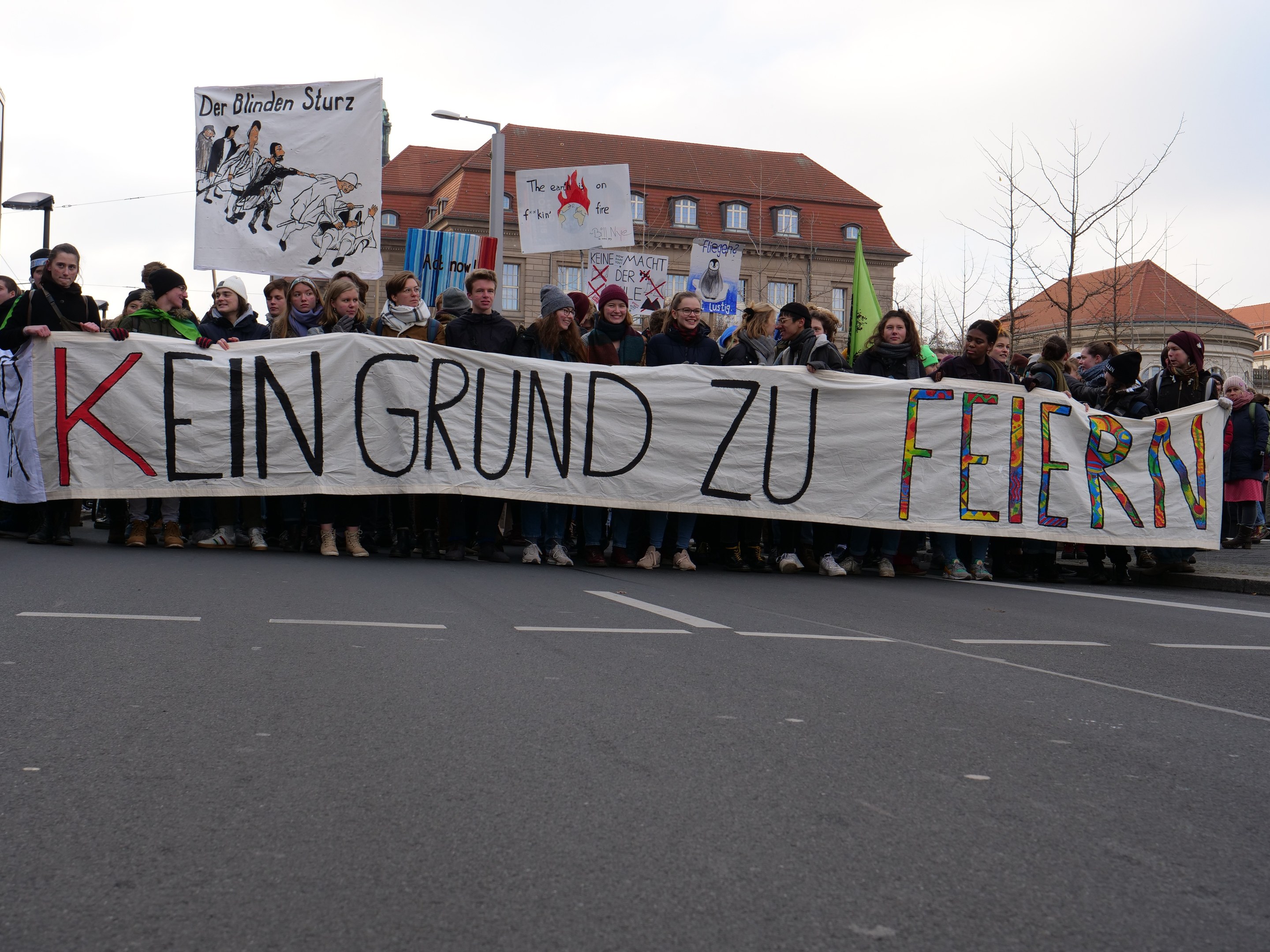 Demonstranten mit einem Transparent mit der Aufschrift 'Kein Grund zur Feier' gegen deutsche Sparmaßnahmen, mit Straßeninfrastruktur und Gebäuden im Hintergrund unter einem klaren Himmel.