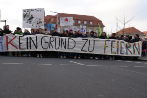 Demonstranten mit einem Transparent mit der Aufschrift 'Kein Grund zur Feier' gegen deutsche Sparmaßnahmen, mit Straßeninfrastruktur und Gebäuden im Hintergrund unter einem klaren Himmel.