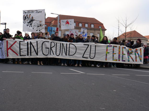Demonstranten mit einem Transparent mit der Aufschrift 'Kein Grund zur Feier' gegen deutsche Sparmaßnahmen, mit Straßeninfrastruktur und Gebäuden im Hintergrund unter einem klaren Himmel.