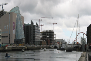 Ein Fluss mit Schiffen, eine Brücke, Gebäude, Kräne und ein Himmel im Hintergrund.